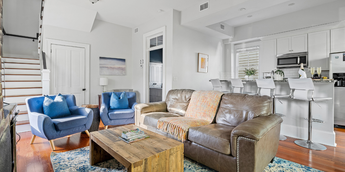 Bright living room with a leather sofa, blue armchairs, and a wooden coffee table beside an open kitchen with breakfast-bar seating.