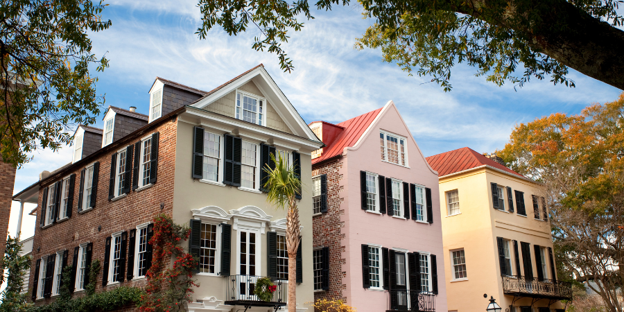 Historic Charleston homes with pastel facades, black shutters, and red roofs beneath leafy trees and a blue sky.