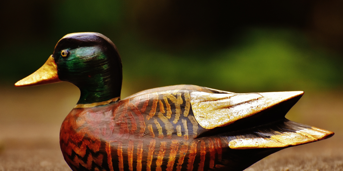 Painted wooden duck decoy with a green head, yellow bill, and patterned brown body against a blurred background.
